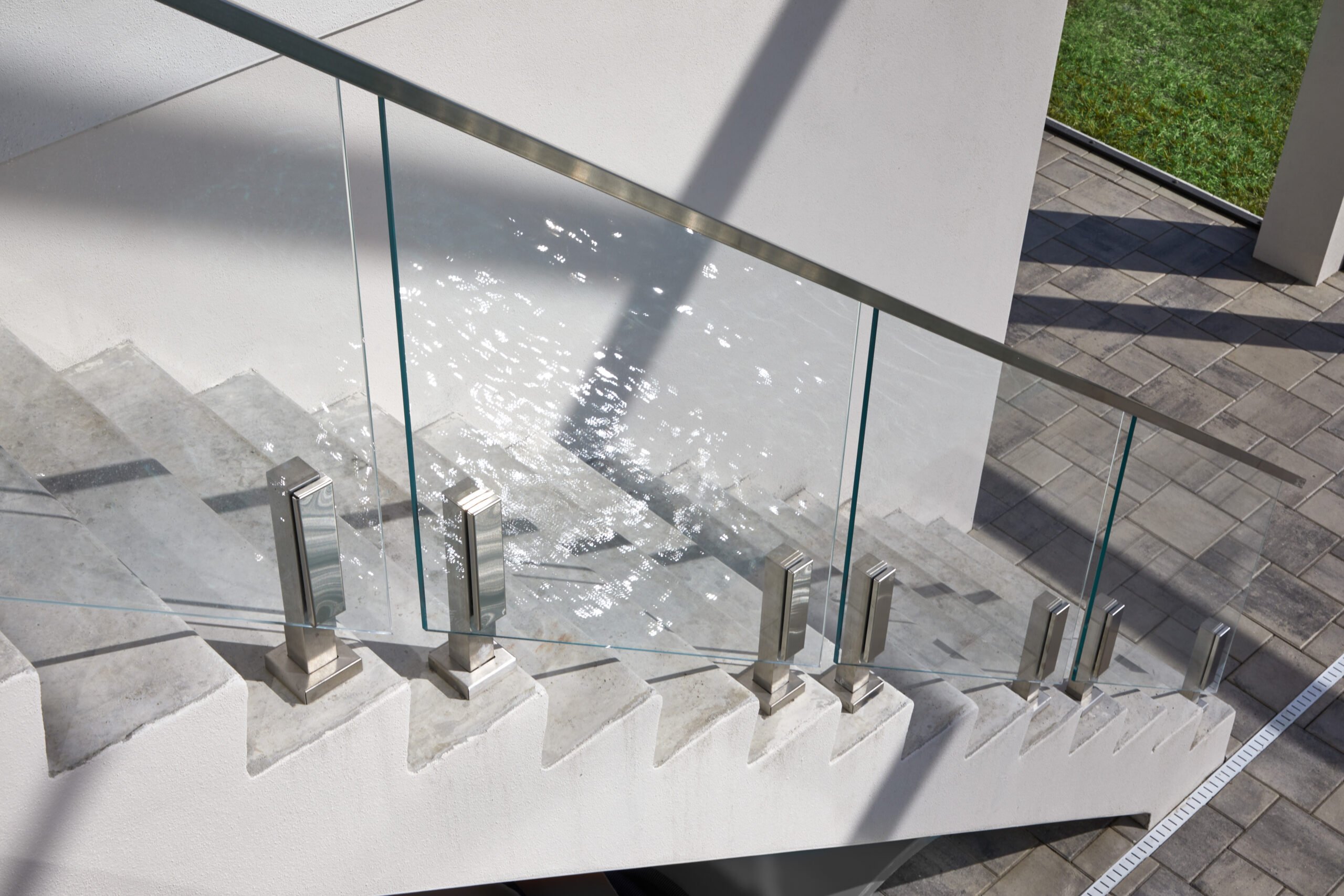 This concrete staircase has a glass railing. The steps lead down to a grey concrete-tiled flooring. There is green grass by the tiled flooring.